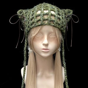 May include: A dark olive green crochet cat-ear hat featuring an open grid pattern, thin brown ribbon bows on each side, and long braided ties hanging down. The hat is crafted from textured yarn and includes small wooden beads at the ends of the braids.