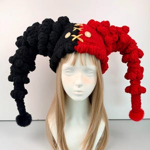 May include: A two-toned jester hat featuring a black side and a red side, both with long, textured, coiled extensions ending in pom-poms. The center front features a gold ribbon lace-up detail secured with two light-colored wooden buttons.