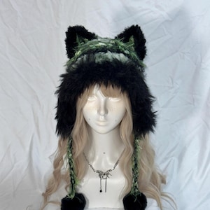 May include: A fuzzy beanie featuring black faux fur trim and ear-like protrusions at the top. The main body of the hat is crafted from a textured green and black yarn blend. Two long braided tassels with black pom-poms hang from the sides of the hat.