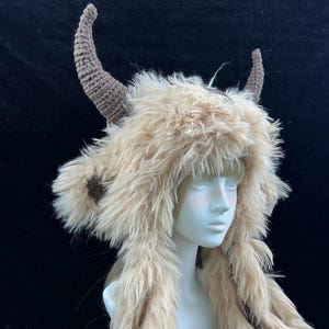 May include: A whimsical hat with a faux fur design and two curved, brown horns. The hat features long, flowing fur extensions on each side. The hat is displayed on a mannequin head against a dark background.