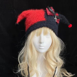 May include: A fuzzy jester-style hat featuring a split red and black design with two pointed ends. The hat is adorned with a black and red patterned ribbon bow, a silver cross charm, and two long black braided ties ending in red star-shaped accents.