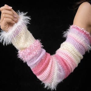 May include: A pair of knitted arm warmers in shades of pink, lavender, and cream. The arm warmers have a striped pattern and are trimmed with fluffy white fringe at the cuffs. The background is solid black.