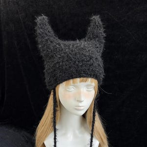 May include: A dark grey, fluffy hat with upright, pointed ears and dangling ties with pom-poms. The hat is shown on a mannequin head with blonde hair, against a black backdrop. The hat's texture looks soft and plush.