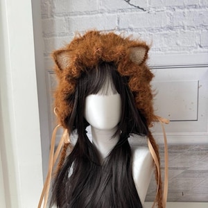 Pode incluir: Um chapéu castanho e peludo com orelhas presas e longas fitas bege. O chapéu foi projetado para se assemelhar a uma juba de leão. O chapéu é exibido em uma cabeça de manequim com cabelo comprido e escuro.