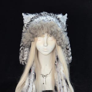 May include: A fluffy, gray and white faux fur hat featuring two small, light-colored wing attachments on the top. The hat has a thick, textured brim and long, dangling side panels made of mixed yarn, fibers, and fur, creating a layered appearance.
