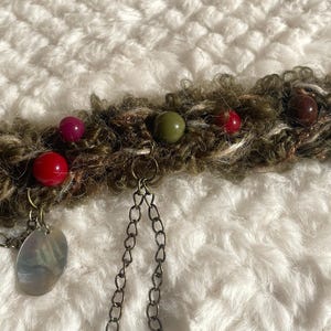 May include: A handmade, bohemian-style bracelet featuring a textured, olive-green and brown band adorned with colorful beads in red, green, and purple. Dangling chains and a pearlescent charm add detail to this unique accessory.