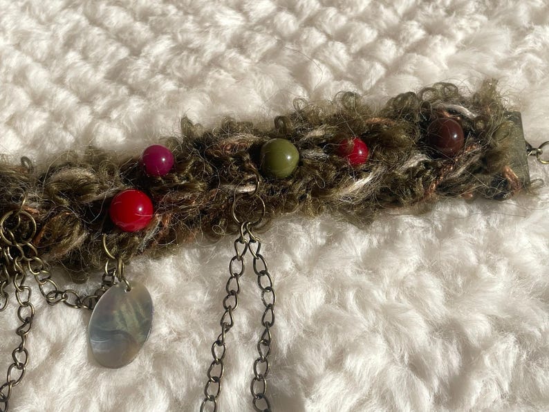 May include: A handmade, bohemian-style bracelet featuring a textured, olive-green and brown band adorned with colorful beads in red, green, and purple. Dangling chains and a pearlescent charm add detail to this unique accessory.