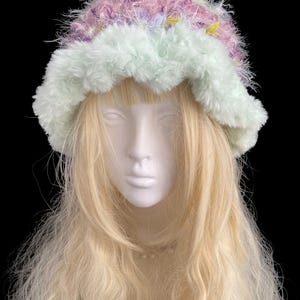 May include: A fluffy, handmade hat with a pastel color scheme. The hat features a light green brim and a crown with pink, purple, and yellow accents. The hat is displayed on a mannequin head with long, blonde hair against a black background.