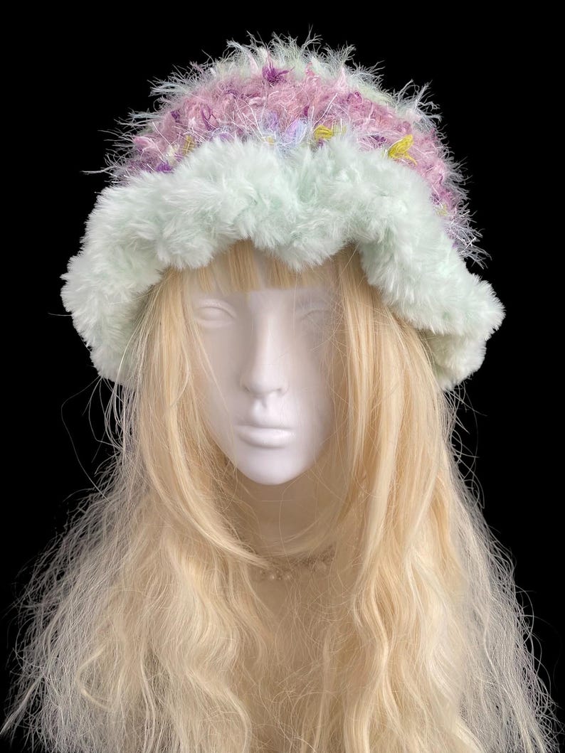 May include: A fluffy, handmade hat with a pastel color scheme. The hat features a light green brim and a crown with pink, purple, and yellow accents. The hat is displayed on a mannequin head with long, blonde hair against a black background.