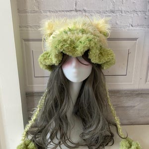 May include: A fuzzy bucket hat in shades of light green and yellow, featuring two pointed ears on top, long braided side ties ending in large pom-poms, and a textured, shaggy surface. The hat is displayed on a mannequin head with long wavy hair.