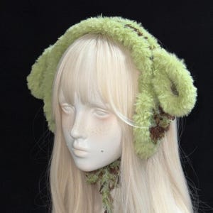 Pode incluir: Uma faixa de cabelo verde claro e fofa com detalhes em marrom. A faixa é projetada com grandes formas semelhantes a orelhas e é feita de um material macio e texturizado. O acessório é exibido em uma cabeça de manequim com cabelos longos e loiros.