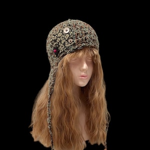 May include: A handmade, crocheted hat in shades of green, brown, and tan, featuring a textured pattern and decorative buttons. The hat has ear flaps and ties, and is displayed on a mannequin head against a black background.