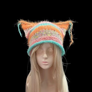 May include: A fuzzy, hand-knitted cat-ear beanie featuring horizontal stripes in vibrant shades of orange, turquoise, and pink. The hat has a textured, loose-knit construction with long, frayed yarn ends extending from the pointed ear tips.