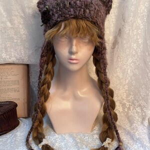 May include: A handmade, purple knit hat with cat ears, adorned with a small flower and ribbon. The hat features long, braided extensions with decorative accents at the ends. The hat is displayed on a mannequin head.