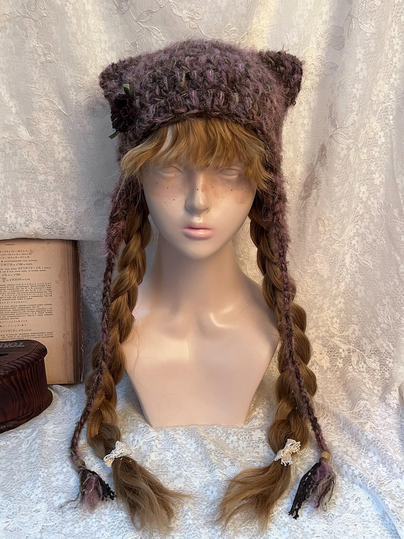 May include: A handmade, purple knit hat with cat ears, adorned with a small flower and ribbon. The hat features long, braided extensions with decorative accents at the ends. The hat is displayed on a mannequin head.