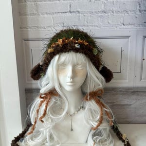 May include: A dark green and brown textured crochet hat with long ear flaps and dangling ties. The hat features a fuzzy, moss-like appearance with small decorative floral accents in shades of yellow, blue, and green attached to the brim and ties.