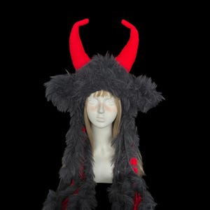 May include: A black and red horned hat with a furry texture. The hat features two large, bright red horns and a long, scarf-like extension with red accents. The hat is displayed on a mannequin head against a black background.