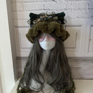 May include: A whimsical olive green faux fur hat with attached ears and a decorative floral and feather trim. The hat is paired with long, wavy gray hair, creating a unique and artistic headpiece.
