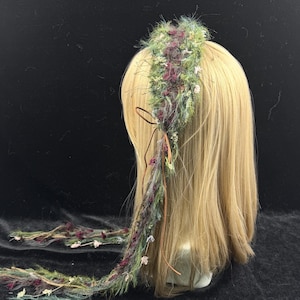 May include: A handmade floral headband with a bohemian style. The headband features a mix of green, purple, and white flowers and foliage. A long, braided strand of the same materials hangs down the back. The headband is displayed on a mannequin head with blonde hair.