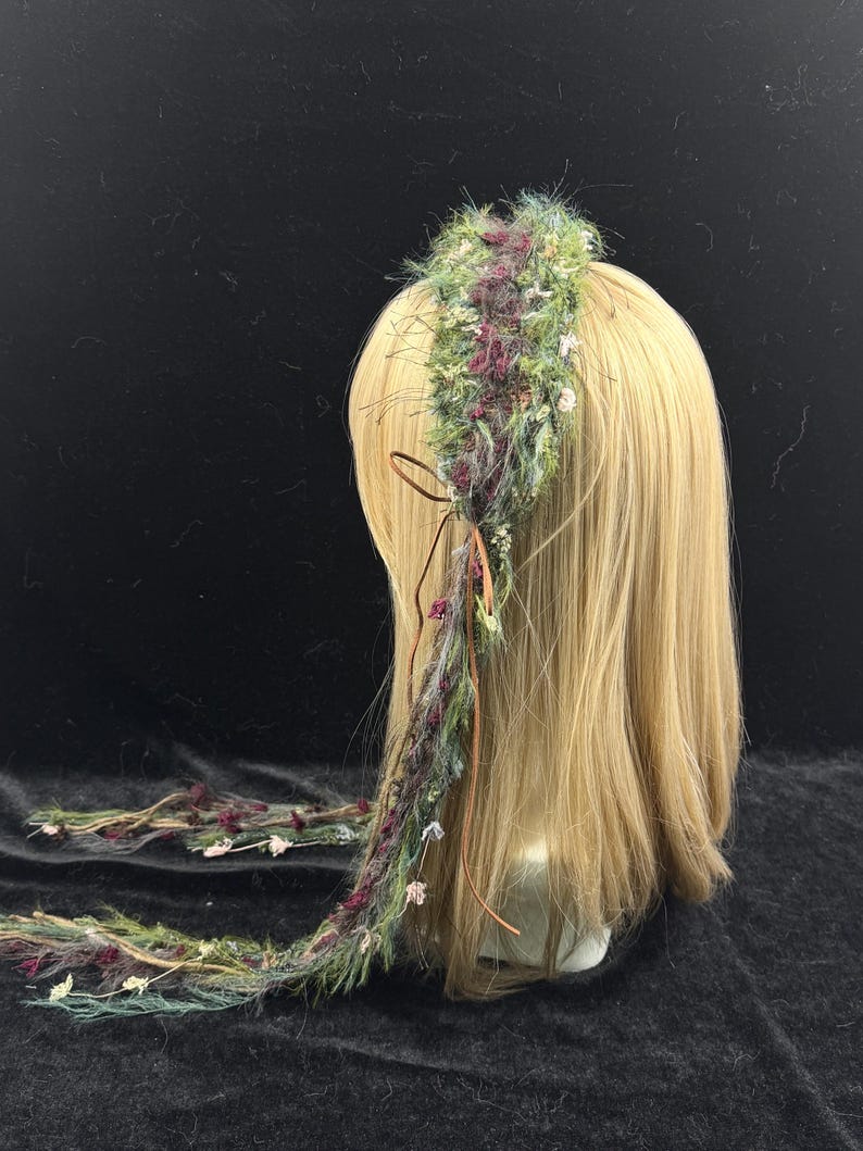 May include: A handmade floral headband with a bohemian style. The headband features a mix of green, purple, and white flowers and foliage. A long, braided strand of the same materials hangs down the back. The headband is displayed on a mannequin head with blonde hair.