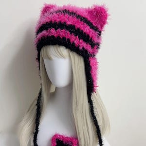 K&ouml;nnte beinhalten: Ein handgefertigter, flauschiger Katzenohrhut in leuchtenden rosa und schwarzen Streifen. Der Hut hat Ohrenklappen und lange B&auml;nder und wird auf einem Schaufensterkopf mit blonden Haaren pr&auml;sentiert. Ein passendes sternf&ouml;rmiges Accessoire ist ebenfalls sichtbar.