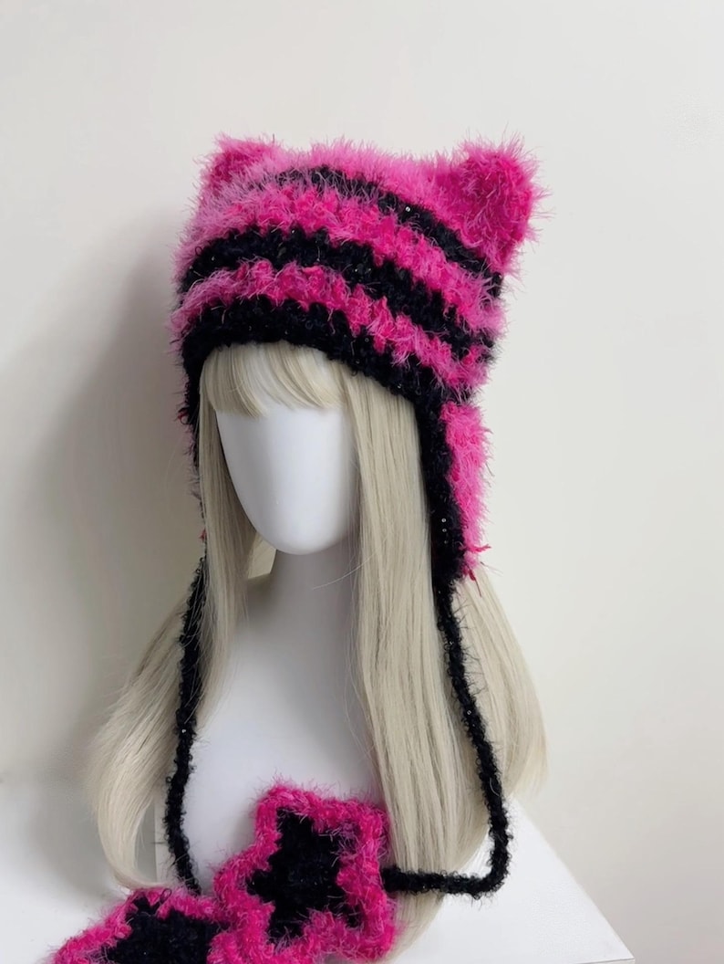 K&ouml;nnte beinhalten: Ein handgefertigter, flauschiger Katzenohrhut in leuchtenden rosa und schwarzen Streifen. Der Hut hat Ohrenklappen und lange B&auml;nder und wird auf einem Schaufensterkopf mit blonden Haaren pr&auml;sentiert. Ein passendes sternf&ouml;rmiges Accessoire ist ebenfalls sichtbar.