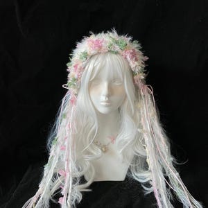 May include: A white wig with long, flowing hair is decorated with a floral crown in pink and green. Strands of white, pink, and green fabric hang down, creating a whimsical and ethereal appearance.