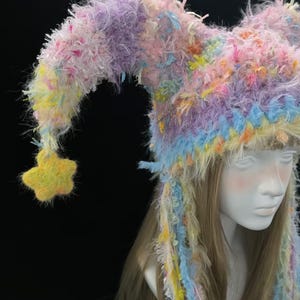 May include: A colourful, whimsical hat with a jester-like design. The hat features a mix of pastel colours, including pink, purple, yellow, and blue. A yellow star hangs from the end of the hat's point. The hat is made of a fuzzy, textured material.