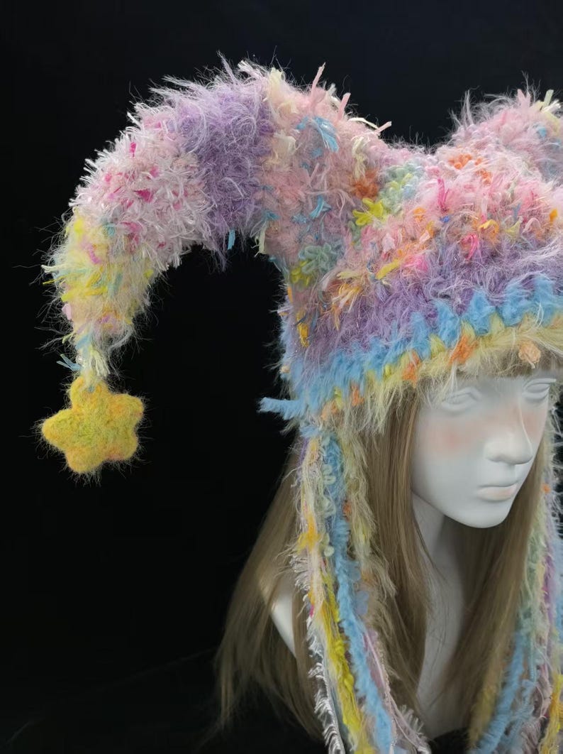 May include: A colourful, whimsical hat with a jester-like design. The hat features a mix of pastel colours, including pink, purple, yellow, and blue. A yellow star hangs from the end of the hat's point. The hat is made of a fuzzy, textured material.