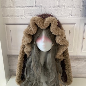 May include: A brown and burgundy faux fur hood with long ear flaps. The hood is lined with a darker burgundy fabric and has a fluffy, textured appearance. The hood is displayed on a mannequin head with long, wavy grey hair.