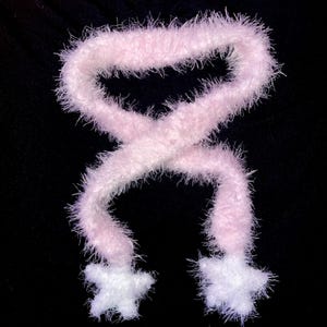 May include: A fluffy, light pink scarf with white star-shaped ends. The scarf is made of a soft, fuzzy material and is displayed against a black background. The scarf is a winter accessory.