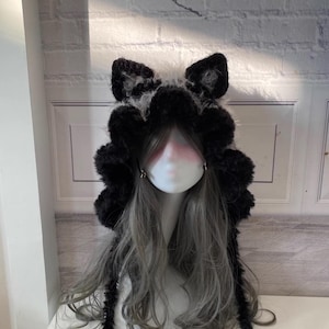 May include: Black faux fur hooded hat with long tails. The hat has cat ears and a fluffy texture.