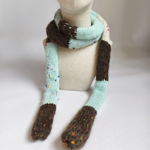 May include: A knitted scarf in brown, pale blue, and white. The scarf features a speckled pattern with colourful flecks. The scarf is displayed on a mannequin head against a white backdrop. A winter accessory.