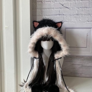 Peut inclure: Un chapeau noir et blanc avec des oreilles de chat, une capuche duveteuse et de longues tresses noires. Le chapeau présente des oreilles intérieures roses et des pompons noirs au bout des tresses. La capuche est bordée de fausse fourrure rose clair.