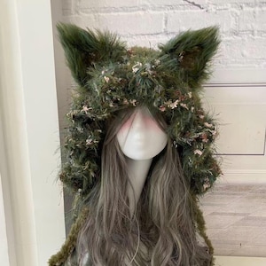 May include: A green faux fur hood with pointed ears, adorned with small pink and white floral accents. The hood is displayed on a mannequin head with long, wavy gray hair. The overall aesthetic suggests a whimsical, fantasy-inspired accessory.