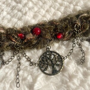 May include: A handcrafted necklace featuring a textured, brown and green yarn base adorned with red beads and metal charms. The necklace includes a tree of life pendant and dangling silver-toned oval charms.