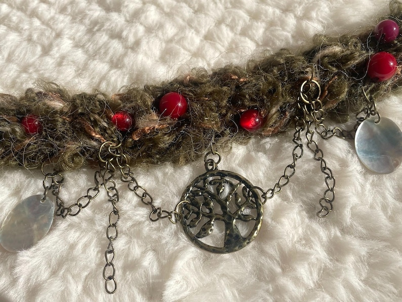 May include: A handcrafted necklace featuring a textured, brown and green yarn base adorned with red beads and metal charms. The necklace includes a tree of life pendant and dangling silver-toned oval charms.