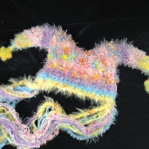 May include: A handmade jester's hat in a vibrant mix of pastel shades. The hat is adorned with pink, yellow, blue, and purple hues. It has two long, flowing tails and yellow star-shaped embellishments at the ends, creating a playful look.