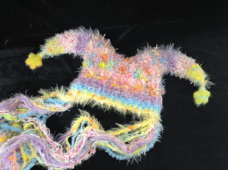 May include: A handmade jester's hat in a vibrant mix of pastel shades. The hat is adorned with pink, yellow, blue, and purple hues. It has two long, flowing tails and yellow star-shaped embellishments at the ends, creating a playful look.