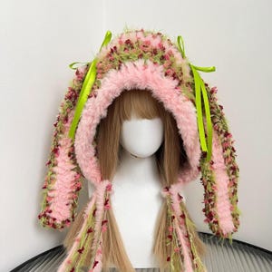 May include: A handmade hood with a pink faux fur lining and exterior. The hood is decorated with green and pink textured details, and lime green ribbons. The hood has long, braided extensions with matching embellishments.