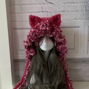 Puede incluir: Una capucha artesanal con forma de orejas de gato, de color magenta, con lazos largos. Está hecha de un material esponjoso y texturizado con detalles en rosa y blanco y pequeñas estrellas. La capucha se muestra en una cabeza de maniquí con pelo largo y ondulado gris.