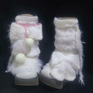 Puede incluir: Un par de botas blancas con plataforma y detalles de punto y pelusa. Una bota presenta una cuerda rosa con dos pequeños pompones, la otra un lazo grande. Las botas tienen una base blanca y una suela de plataforma gris.