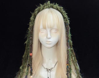 May include: A green textured yarn headpiece featuring long, hanging strands adorned with colorful beads and braided accents, worn on a pale mannequin head with long blonde hair and a silver butterfly-shaped pendant necklace.