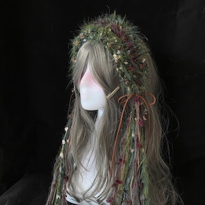 May include: A gray wig with long, wavy hair, adorned with a green and burgundy floral crown. Strands of colorful yarn and dried flowers cascade down the front. A gold hair clip and brown leather ties add detail.