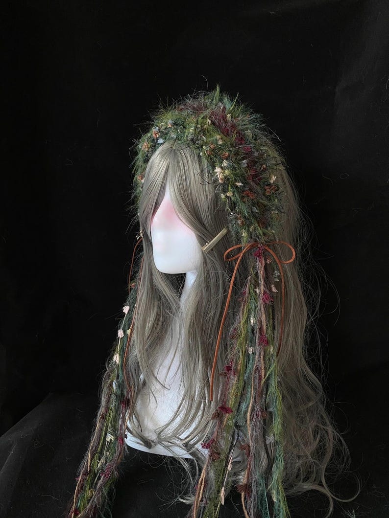 May include: A gray wig with long, wavy hair, adorned with a green and burgundy floral crown. Strands of colorful yarn and dried flowers cascade down the front. A gold hair clip and brown leather ties add detail.