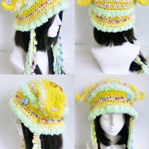 May include: A handmade yellow crochet hat with a whimsical design. The hat features a ruffled brim, pastel accents, and dangling beaded strands. The hat is adorned with decorative buttons and a bow.