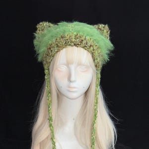 Puede incluir: Un gorro de punto verde y marrón con orejas de gato y largas cintas trenzadas. El gorro presenta un ribete esponjoso de color verde claro y un diseño de punto texturizado. El gorro se muestra en una cabeza de maniquí con cabello rubio.