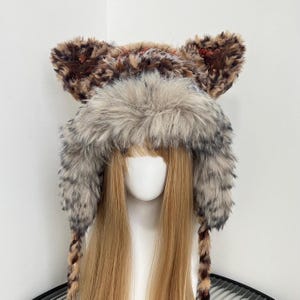 May include: A faux fur hat with attached ears, featuring a mix of brown, gray, and tan colors. The hat has a fluffy texture and long braids. The ears are a darker brown with a spotted pattern. The hat is designed to be worn on the head.
