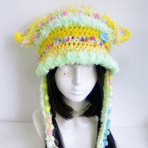 May include: A handmade, colorful crochet hat with a whimsical design. The hat features a yellow base with stripes of pastel green, pink, and white. It has ear flaps and dangling strands with pom-poms. A blue heart-shaped button adorns the side.