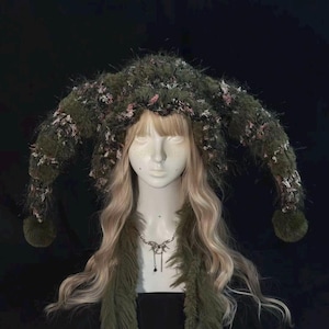 May include: A dark olive green knit hat with long, curved, horn-like side extensions featuring a fuzzy, textured yarn blend with flecks of pink and white. The hat has a pom-pom at each end and is displayed on a mannequin head with long blonde hair.